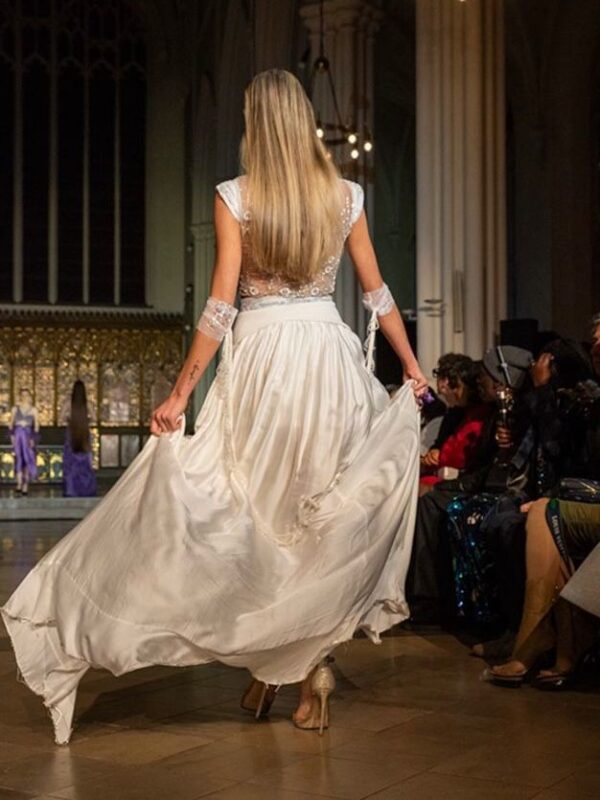 Model walking the runway at London Fashion Week wearing an ivory hand-embroidered blouse with a flowing satin draped skirt by Saanjh by Saloni. Back view: Back view of ivory draped satin skirt in motion with detailed hand-embroidered blouse at London Fashion Week.